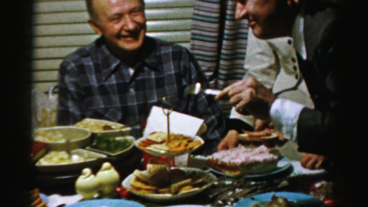 A smiling man sits at a food-filled table as other adults gesture and hold small plates nearby.