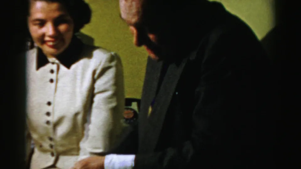 A woman in a cream buttoned jacket smiles beside a man in a dark coat inside a 1950s Albany private home interior.
