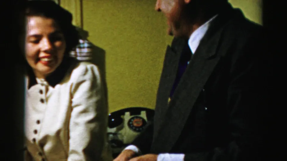 In a 1950s Albany home interior, a smiling woman sits near a man in a dark coat while a black rotary telephone rests behind them.