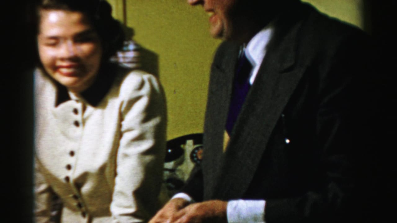 In supported Albany 1950s home-movie footage, a woman smiles beside a cropped suited man in a warmly lit room.