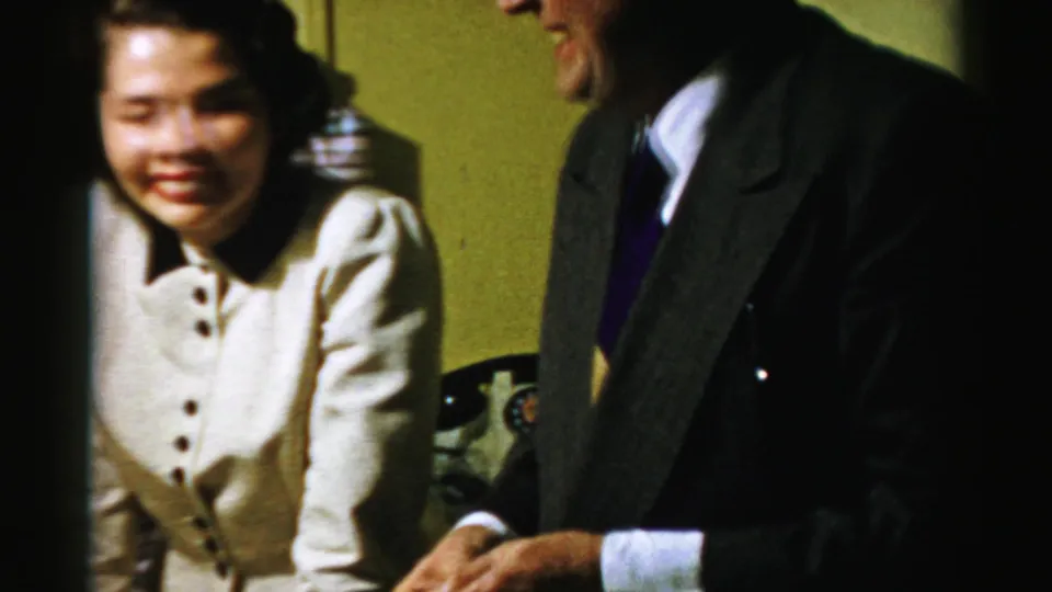 In supported Albany 1950s home-movie footage, a woman smiles beside a cropped suited man in a warmly lit room.