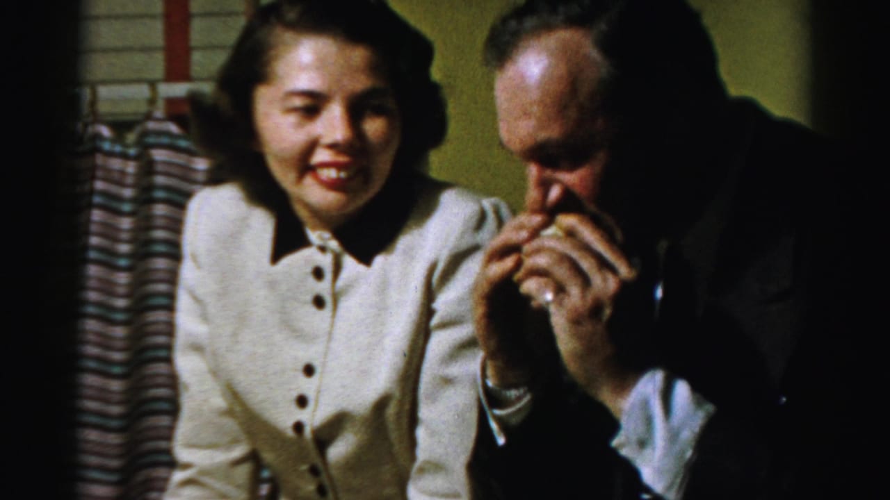 Woman smiles while a suited man raises a sandwich near his mouth indoors.