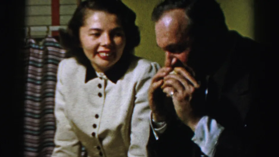 A woman watches with amusement as a suited man brings a sandwich to his mouth in supported Albany 1950s home-movie footage.