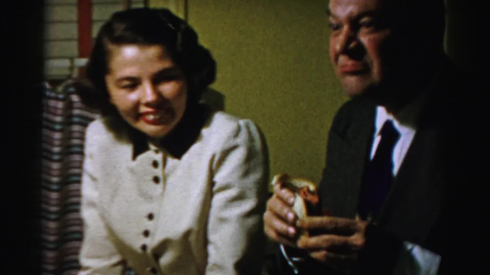 In the Albany 1950s home-movie scene, the man holds a sandwich with a comic expression while the woman smiles nearby.