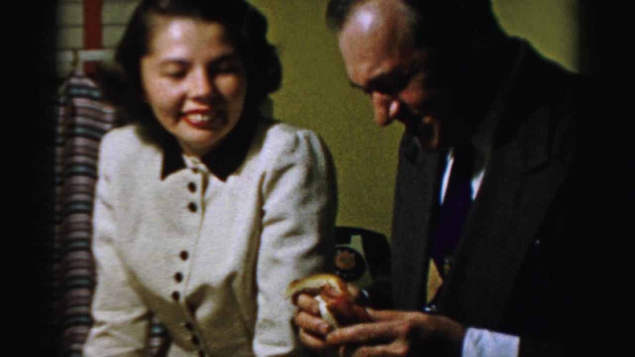 In supported 1950s Albany home-movie footage, a seated woman smiles as a suited man looks down at food in a warm private room.