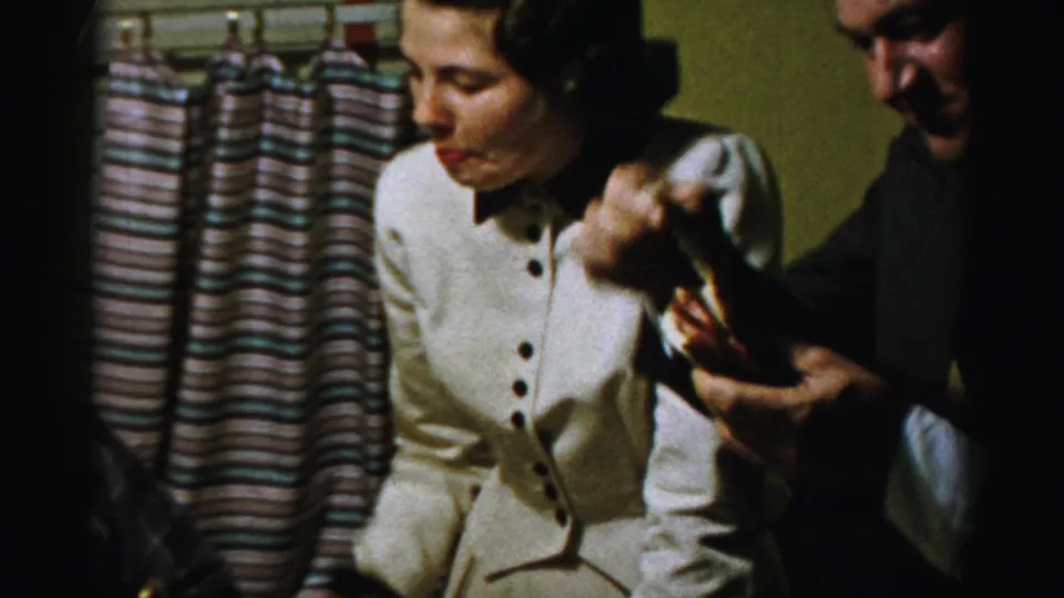 A seated woman in a light dress watches as a suited man holds up a small piece of food inside a mid-century private room.