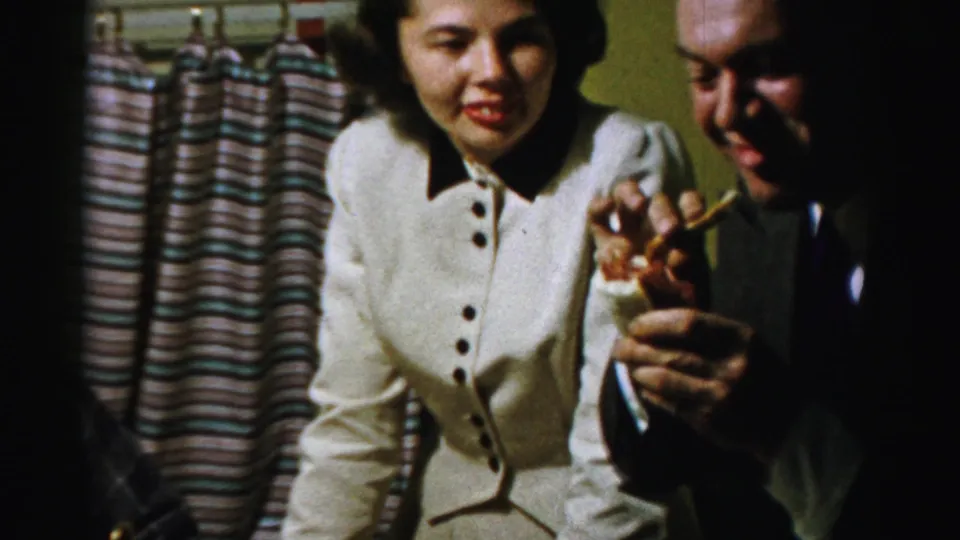 The woman smiles toward the camera while the suited man laughs and holds a small food item in this 8mm domestic scene.