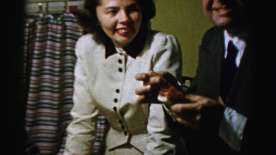 A smiling woman sits beside a partly cropped suited man holding food, with striped curtains behind them in a private Albany interior.