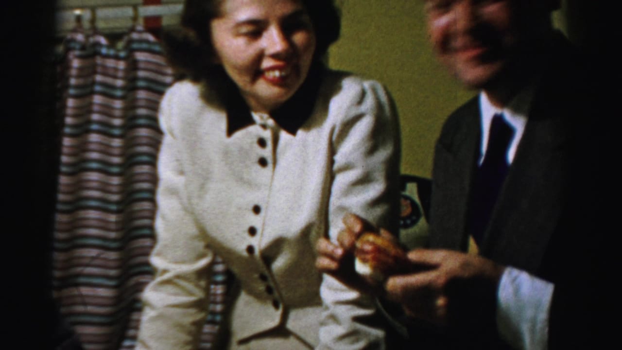 In 1950s Albany home-movie footage, a woman in a cream jacket smiles while seated beside a man in a suit during an indoor meal.