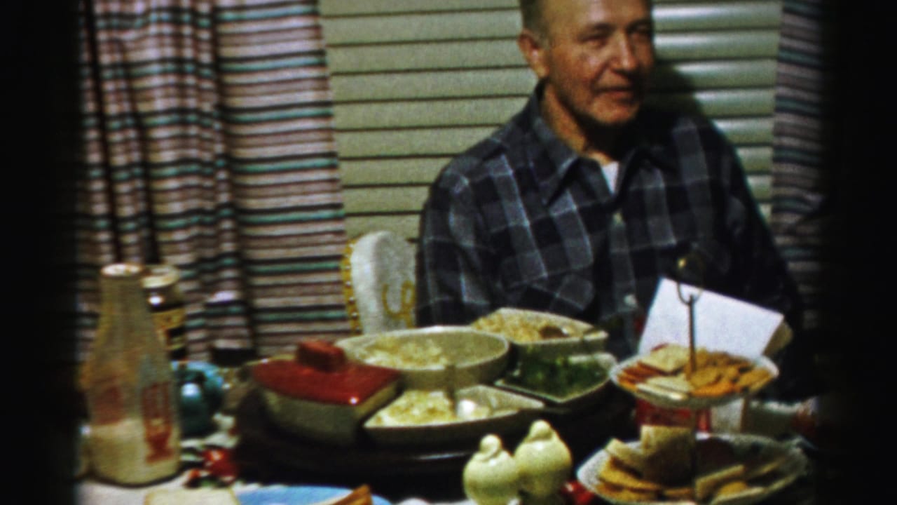 Man in a plaid shirt sits behind a table filled with dishes, snacks, cups, and a glass bottle.