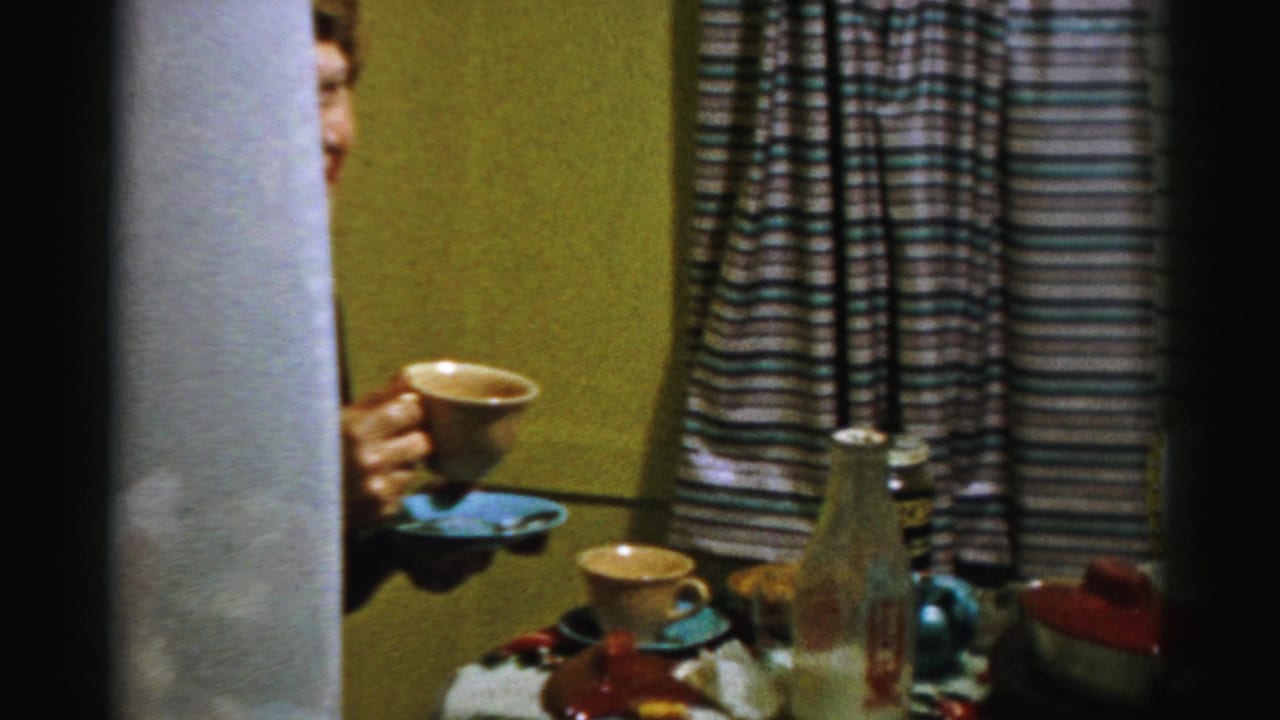 In Albany home-movie footage from 1956, a partially visible older woman holds coffee near a table with cups, dishes, and striped curtains.