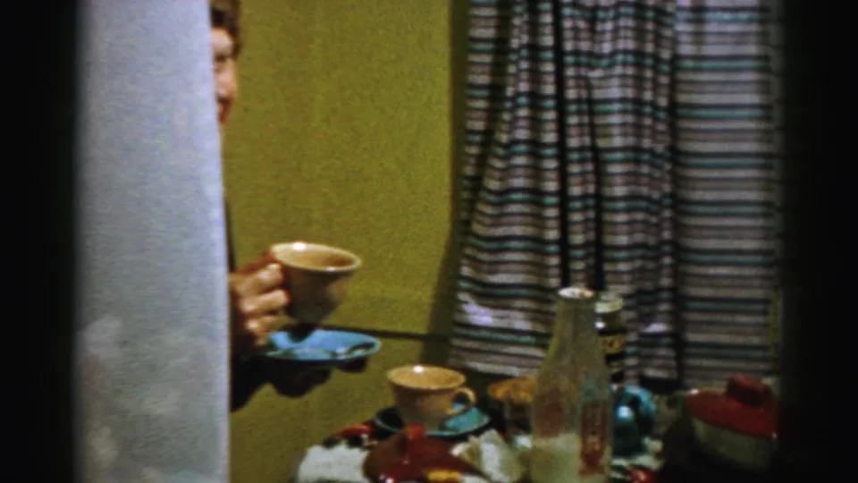 In Albany home-movie footage from 1956, a partially visible older woman holds coffee near a table with cups, dishes, and striped curtains.