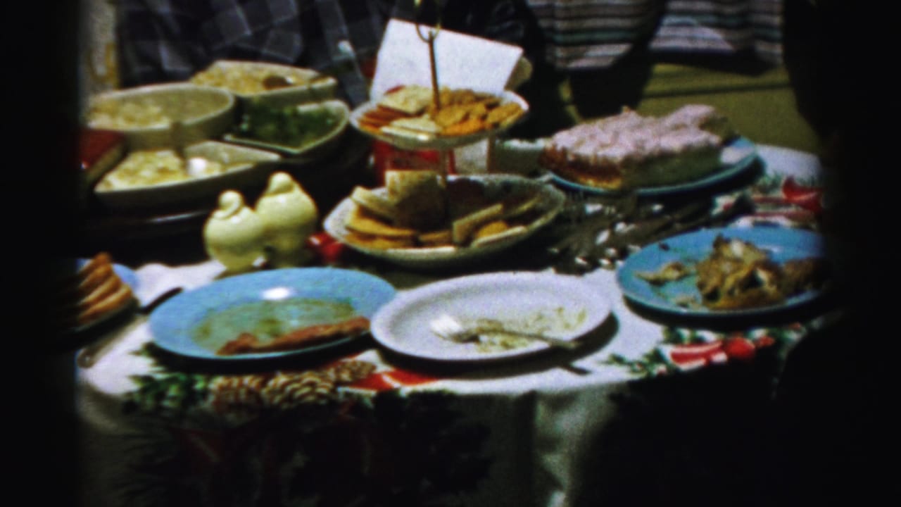 1956 Albany home-movie frame showing a Christmas tablecloth, used plates, bread, cake, serving bowls, and ceramic bird shakers.