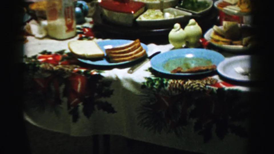 1956 Albany home-movie frame showing bread slices, a pitcher, serving dishes, used plates, and bird-shaped shakers on a Christmas tablecloth.