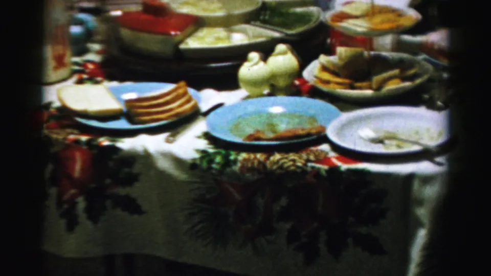 1956 Albany home-movie frame of a Christmas meal table with bread, used plates, bowls, ceramic bird shakers, and greenery along the cloth edge.
