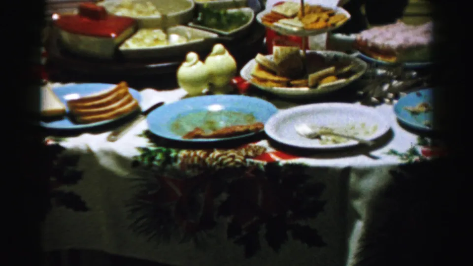1956 Albany home-movie frame of a holiday table with blue plates, bread, serving dishes, and a holly-patterned cloth.