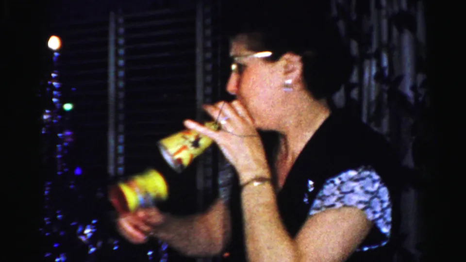 1956 Albany home-movie frame showing an adult woman drinking indoors, with blinds and nighttime city lights in the background.