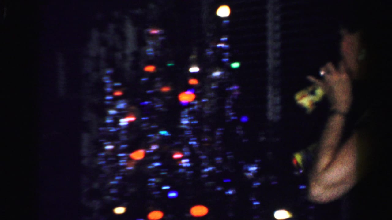 In Albany, New York home-movie footage, a woman stands near a dark window with colorful city lights visible outside.