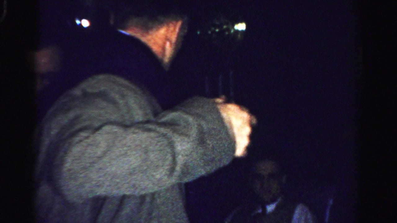 Two adult men appear in a dim Albany holiday interior, one in a gray coat and one in a dark coat, with another figure partly visible.