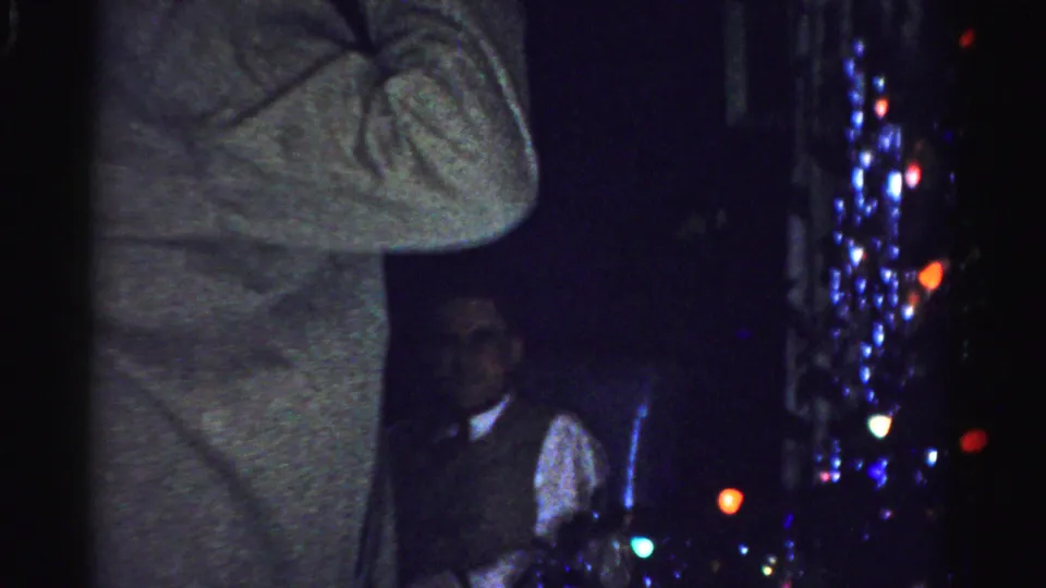 A man in a gray coat stands in front of colored holiday lights, with a seated man in vest and tie partly visible behind him.