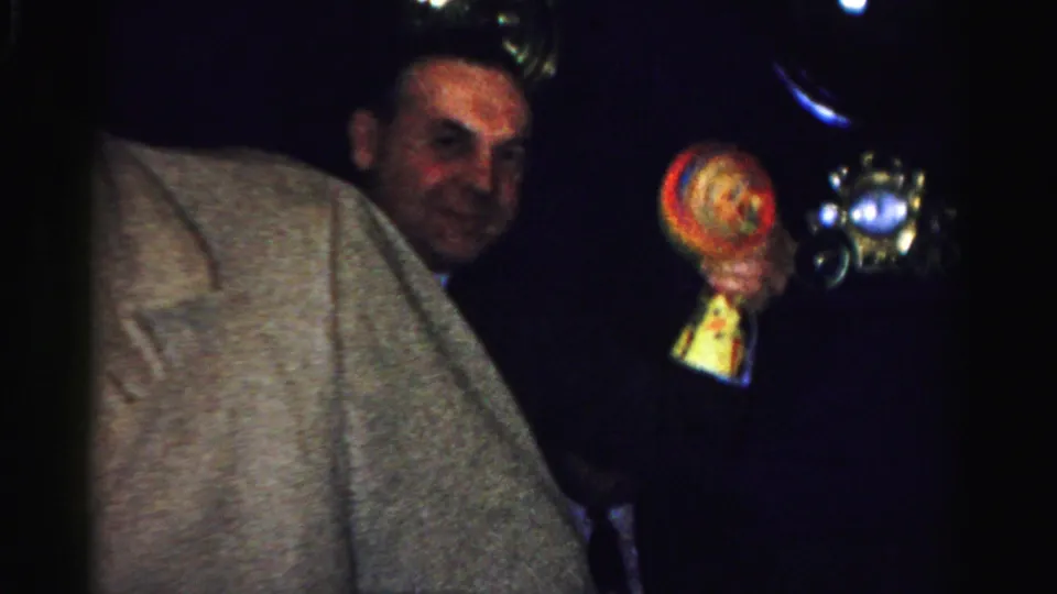 A smiling man in a dark coat holds a small colorful object near another adult in a light coat during the indoor gathering.