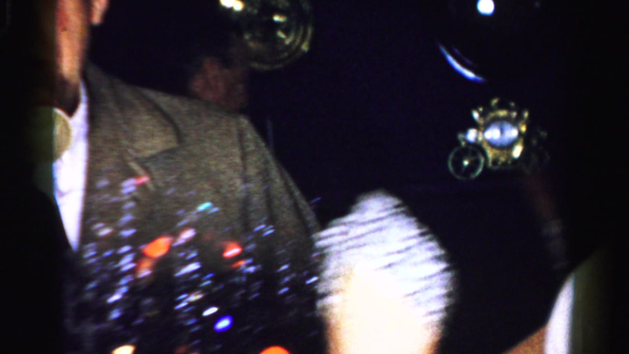 Dim party scene with a man in a tweed jacket beside colored lights and blurred figures.