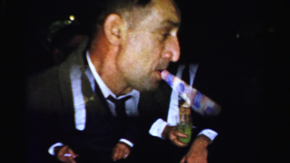 Albany 1950s home-movie frame showing an older suited man with a party blower, drink, and cigarette in a dim party setting.