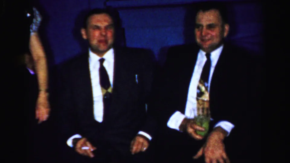 Albany 1950s home-movie frame showing two seated suited men with cigarettes and drinks during an indoor New Year's gathering.