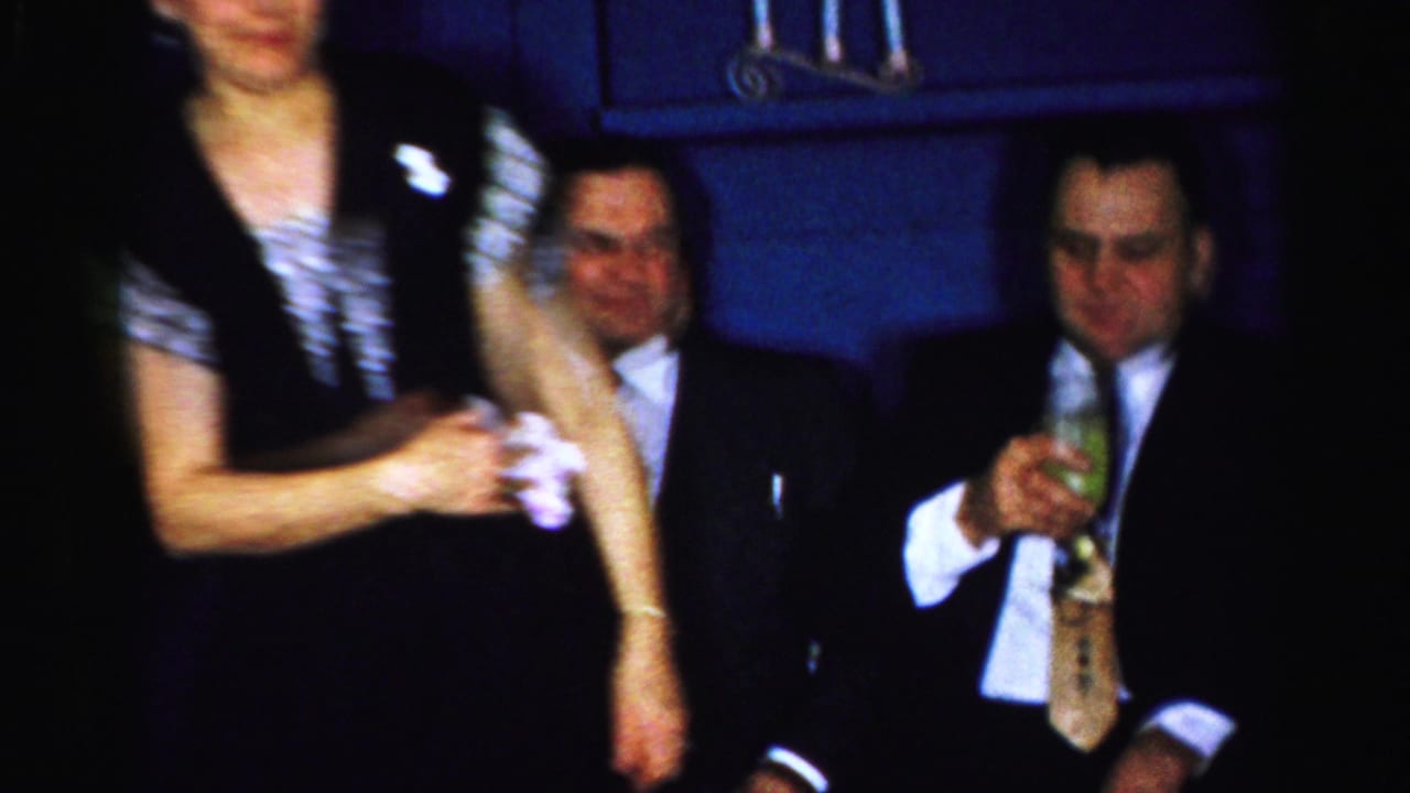 In Albany 1956 home-movie footage, a woman stands near two seated men in suits, one holding a drink, inside a dark room.