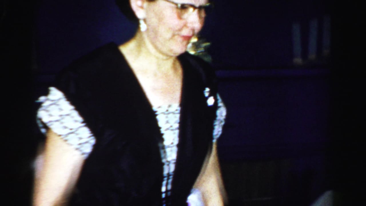An older woman with glasses and a black vest or dress walks through the frame during an Albany 1956 indoor social gathering.
