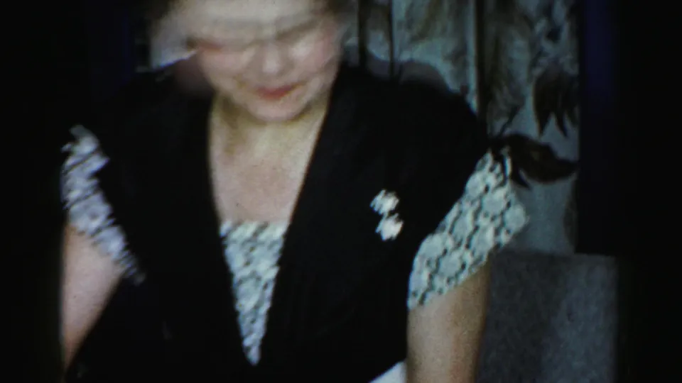 The woman in glasses looks downward in a dim Albany 1956 interior, with dark clothing, patterned sleeves, and household furnishings nearby.