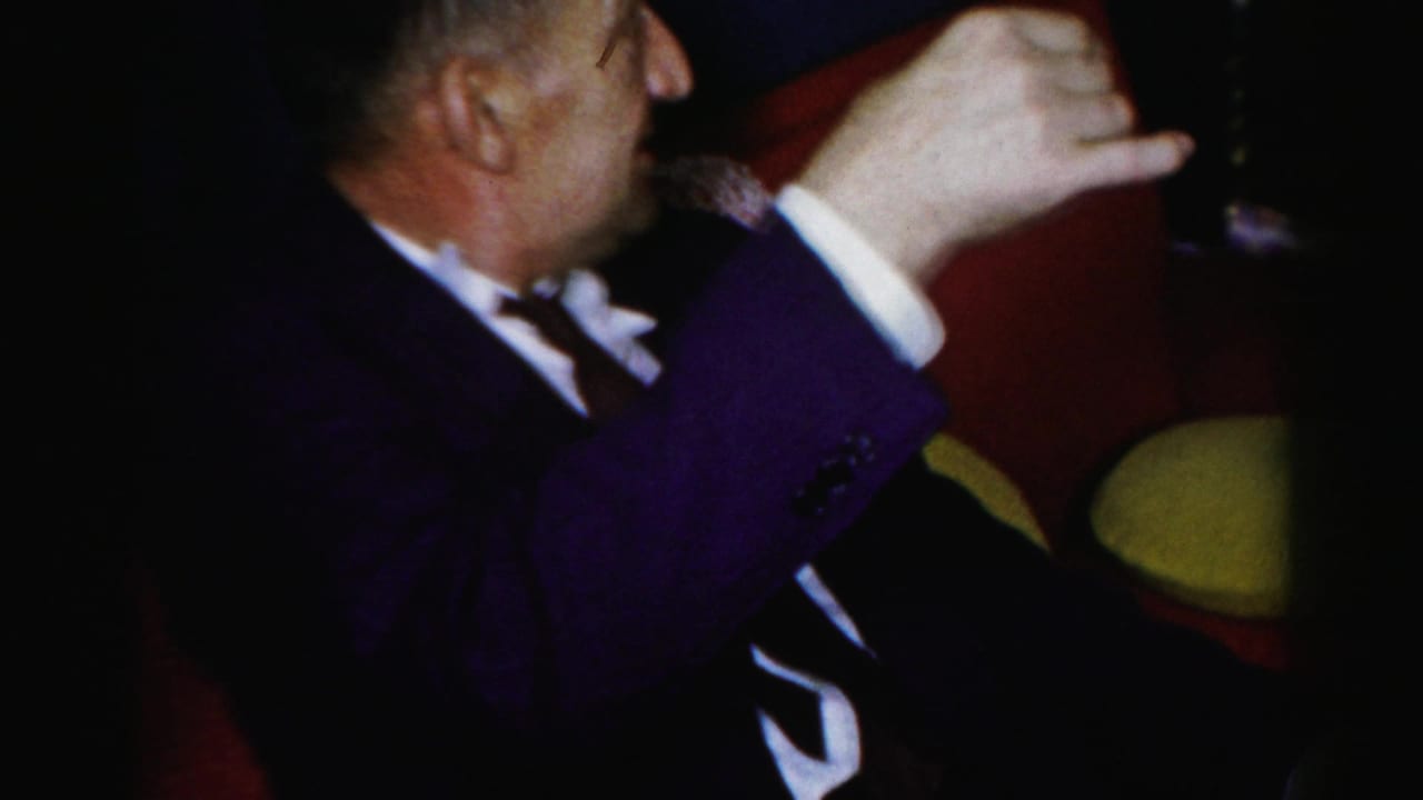 A seated man in a dark suit raises a glass near red upholstered chairs.
