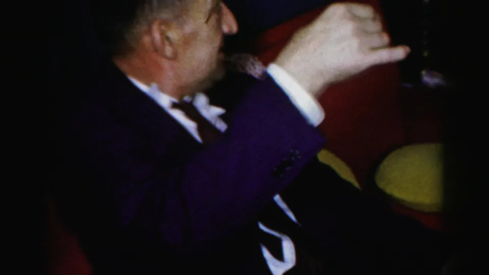 A man in formal dress raises a drink while seated in a red upholstered chair during the Albany gathering.