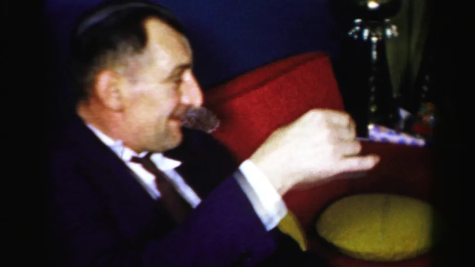 The seated man smiles after gesturing with his drink in the private Albany gathering, with red and yellow chairs behind him.