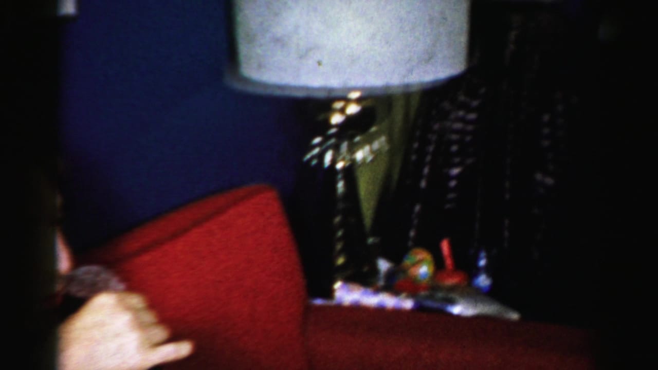 Man seated on a red couch in a dim living room beside a white-shaded lamp.