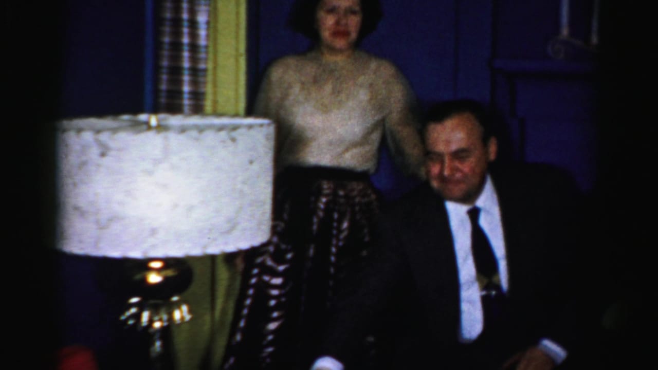 Woman standing behind a seated man in a suit near a lamp and dark chairs.