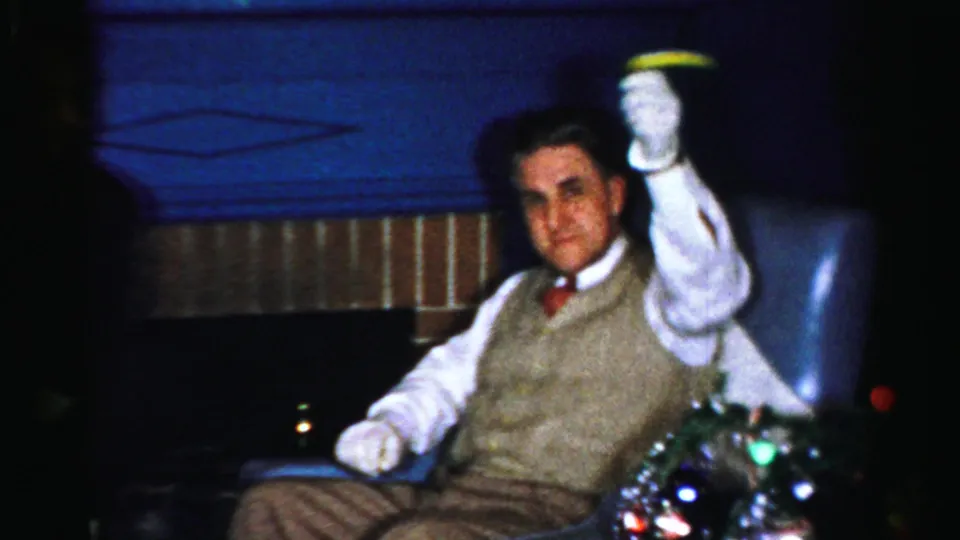 1950s Albany home-movie frame of a seated man in a sweater vest raising a drink near holiday lights.