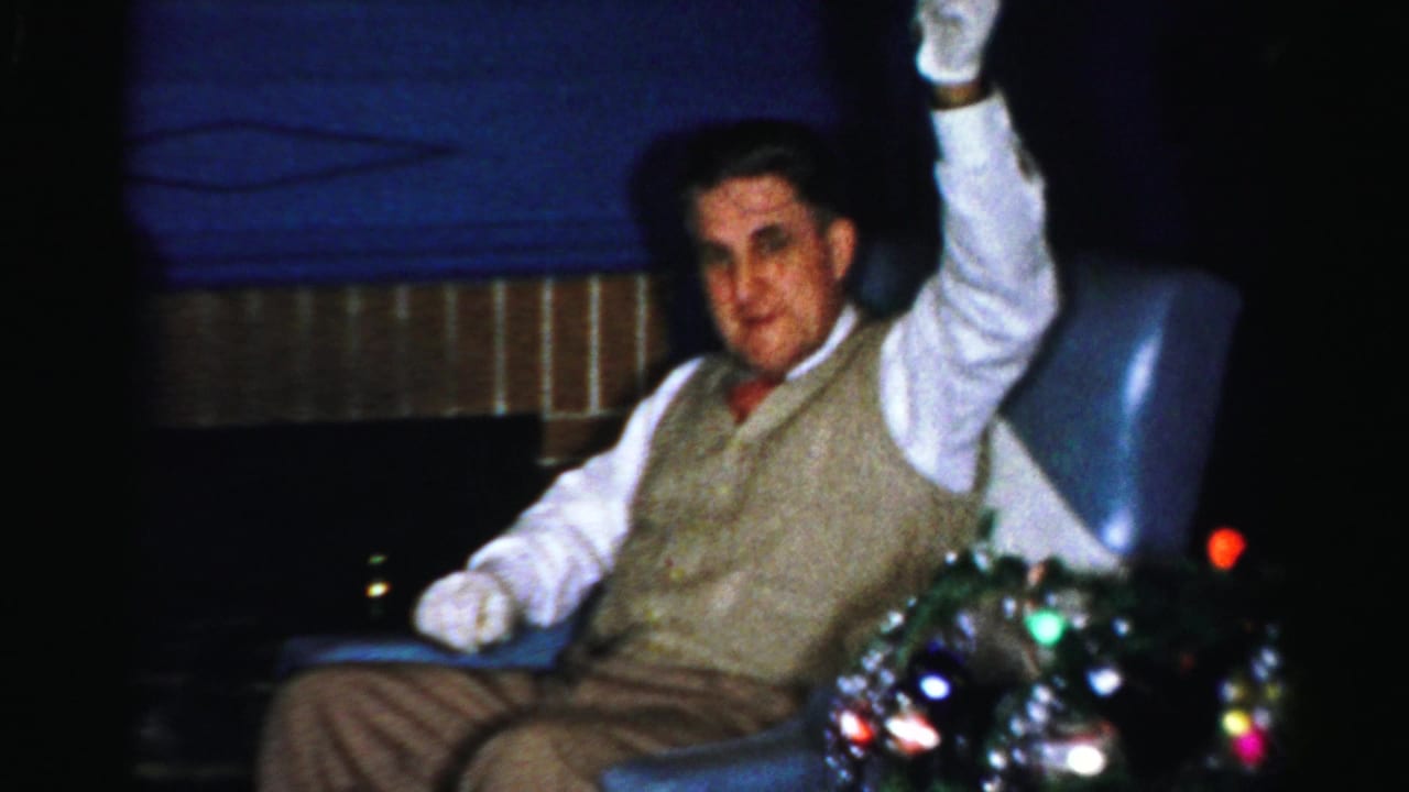 Seated man in vest and white gloves raises a bell beside blue drapery and party lights.
