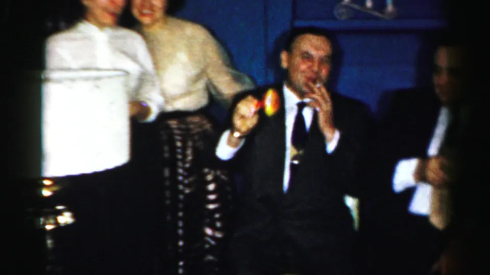 A tuxedoed man holds a bell near two women during a festive 1950s Albany home-movie party moment.