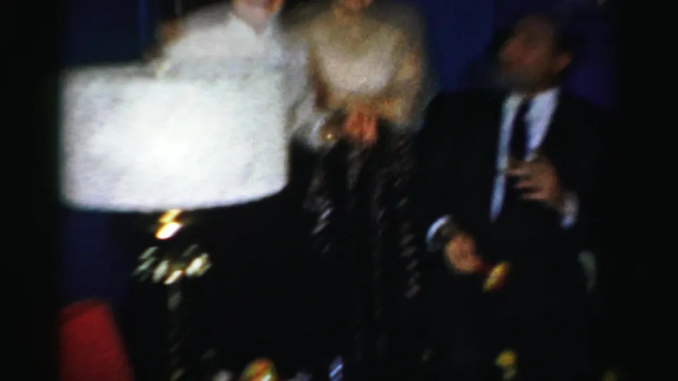 Motion-blurred guests pass through a dim Albany party scene while a suited man holds a drink in 1950s home-movie footage.