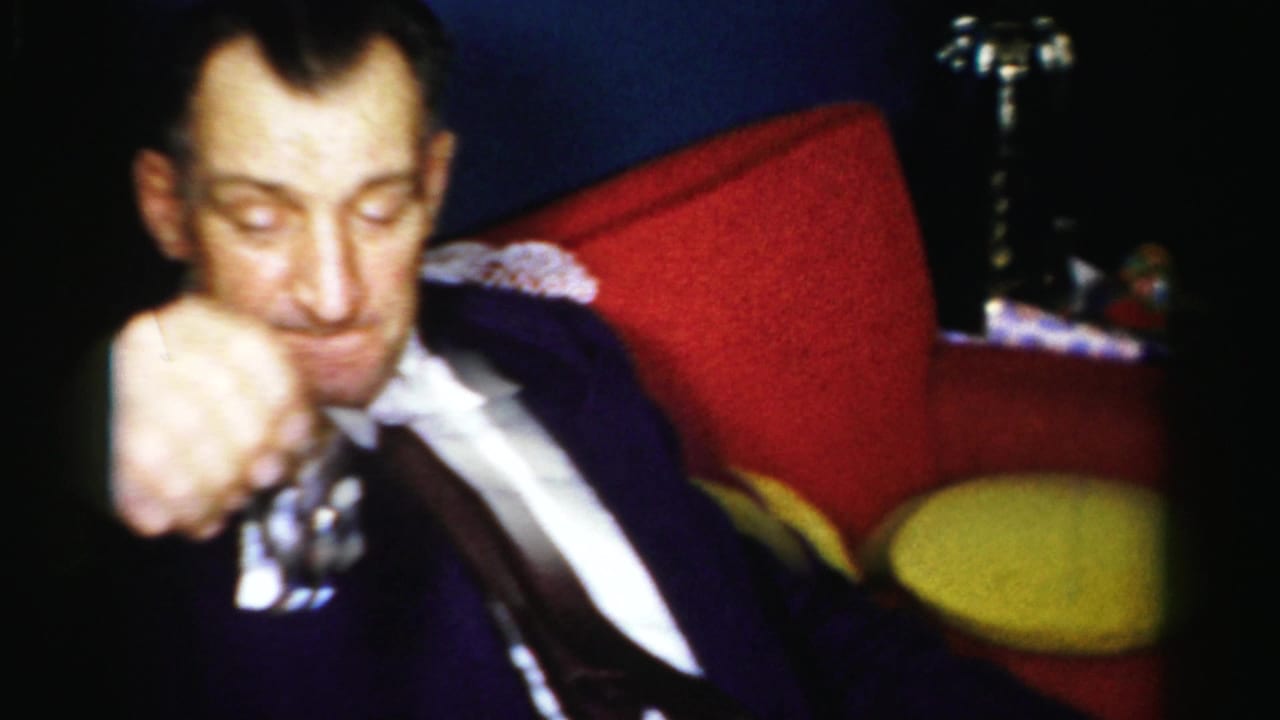 In 1950s Albany home-movie footage, a man in a dark suit sits on a red couch in a dim private interior.