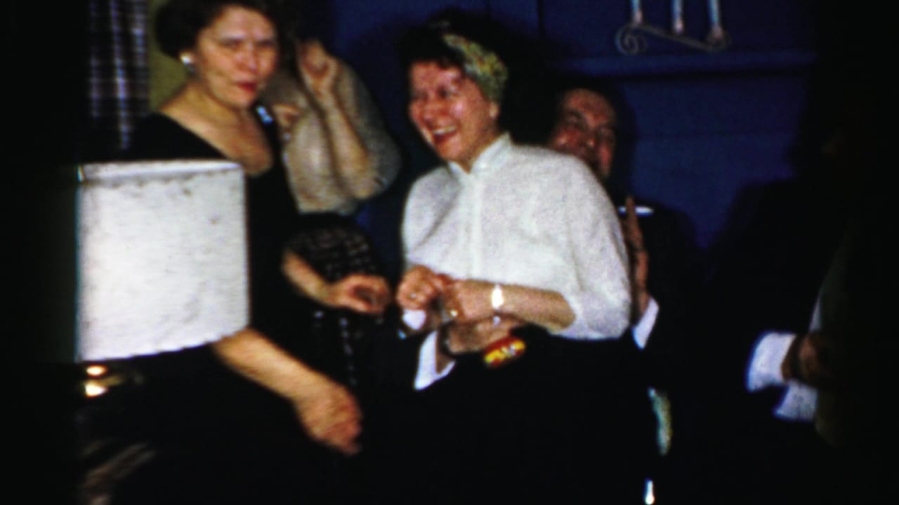 Adults in suits and dresses laugh and hold drinks in a dim Albany private interior from the supplied 1950s home movie.
