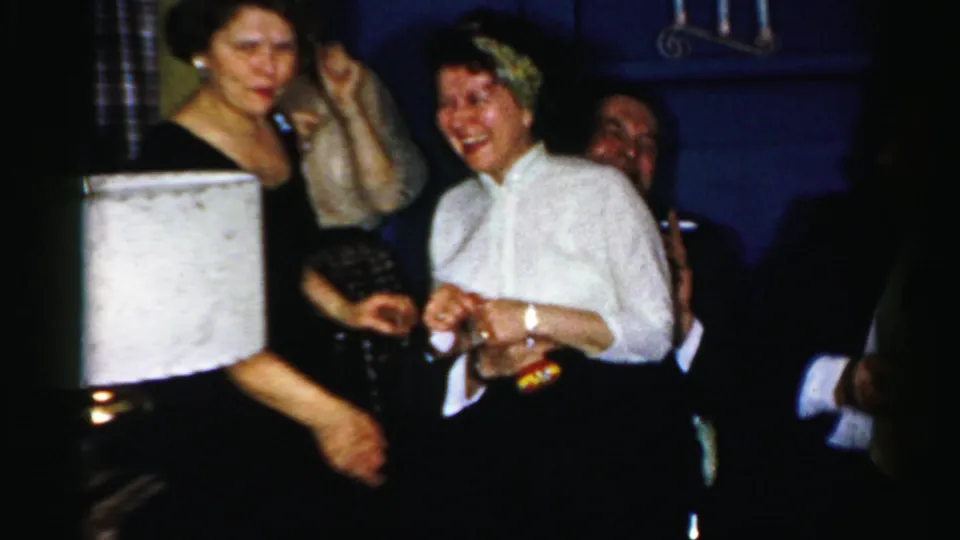 Adults in suits and dresses laugh and hold drinks in a dim Albany private interior from the supplied 1950s home movie.