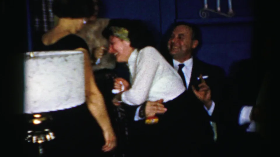 Several adults lean close and laugh while holding drinks beside a lamp in the 1950s Albany home-movie party scene.