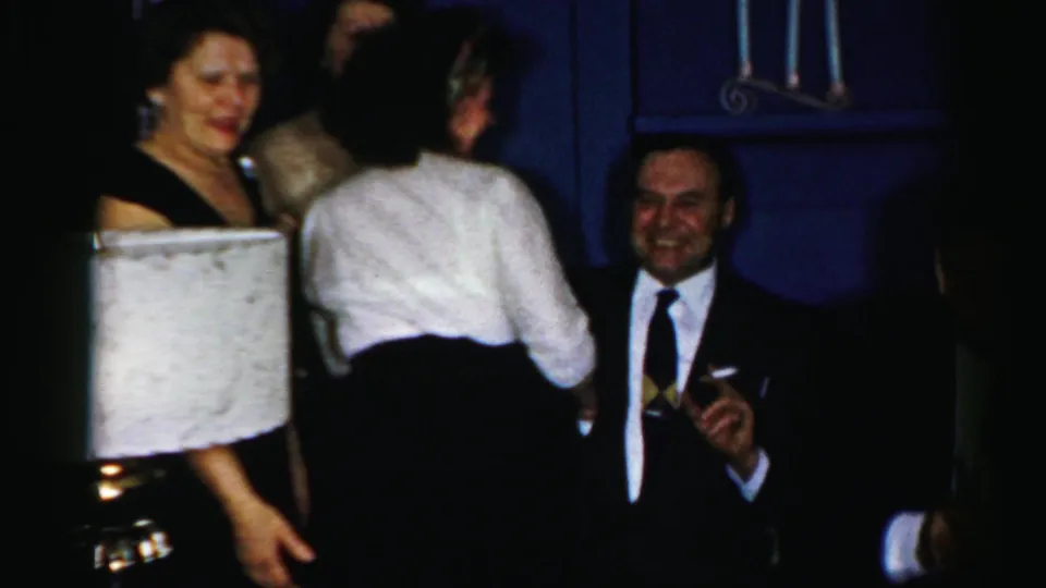 A woman stands with her back to camera as a suited man smiles near a lamp in the 1950s Albany private party footage.