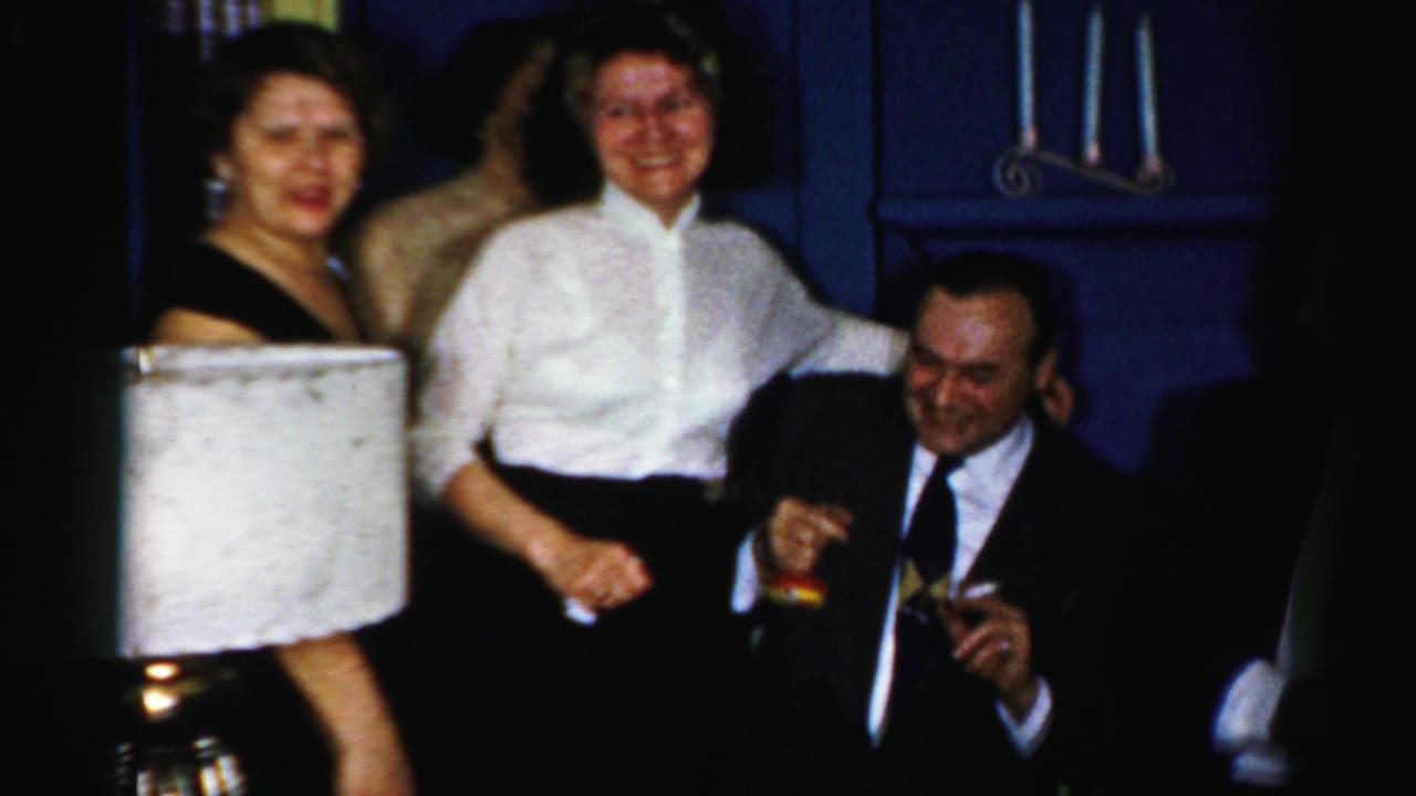 1950s Albany home-movie frame of adults at an indoor gathering, including women in dressy tops and a seated man with a drink.