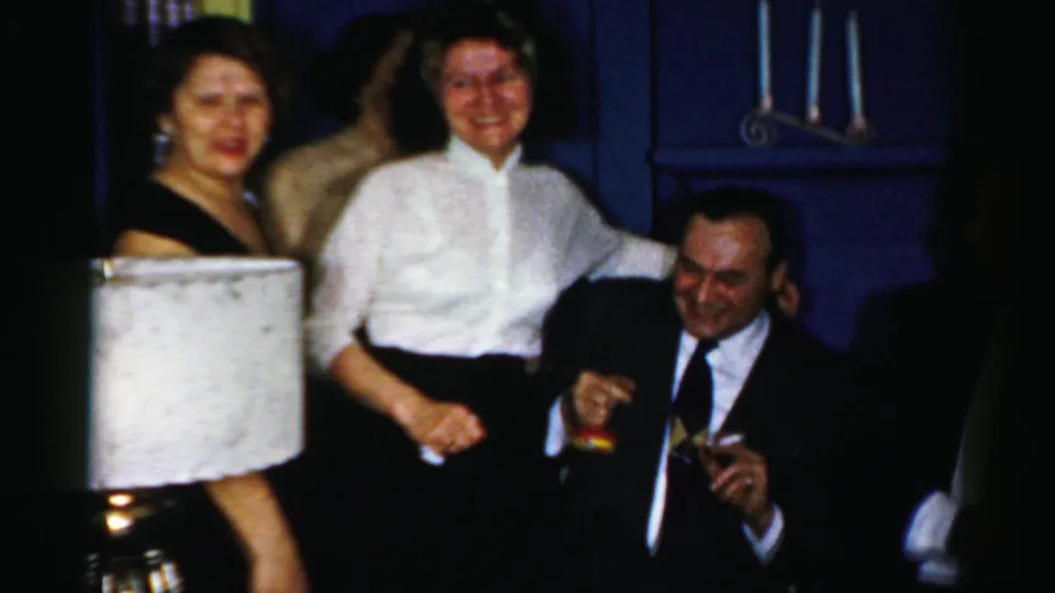 1950s Albany home-movie frame of adults at an indoor gathering, including women in dressy tops and a seated man with a drink.