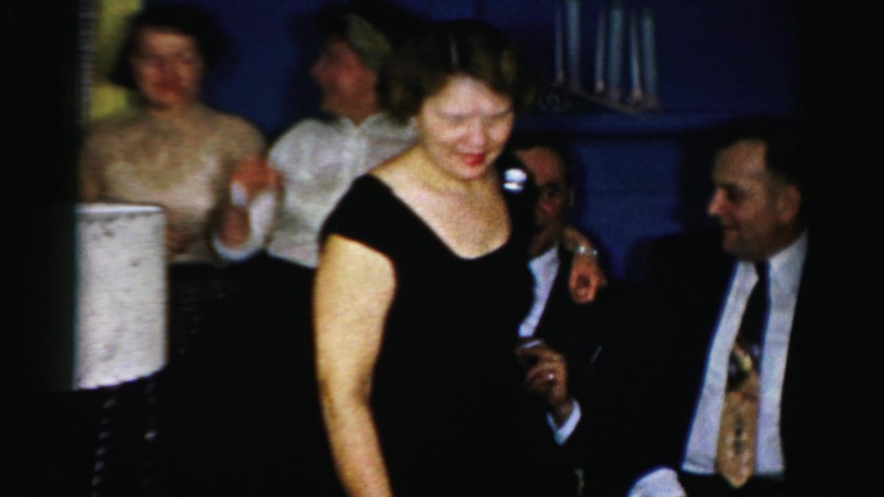 1950s Albany home-movie frame showing a woman in a black dress passing in front of seated adults at a private gathering.