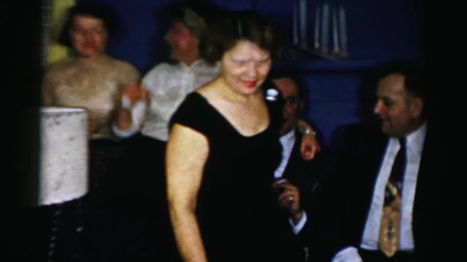 1950s Albany home-movie frame showing a woman in a black dress passing in front of seated adults at a private gathering.