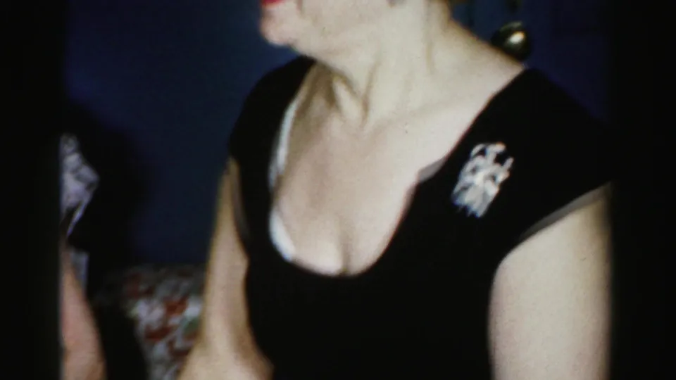 1950s Albany home-movie frame with a cropped close view of a woman in a black dress and brooch inside a private room.